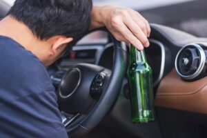 Driver holding a bottle of beer while operating a car, symbolizing the dangers of driving under the influence (DUI) and the importance of not drinking and driving.