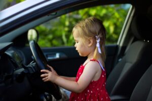 A young girl happily pretends to drive, playing with the steering wheel inside a parked car.